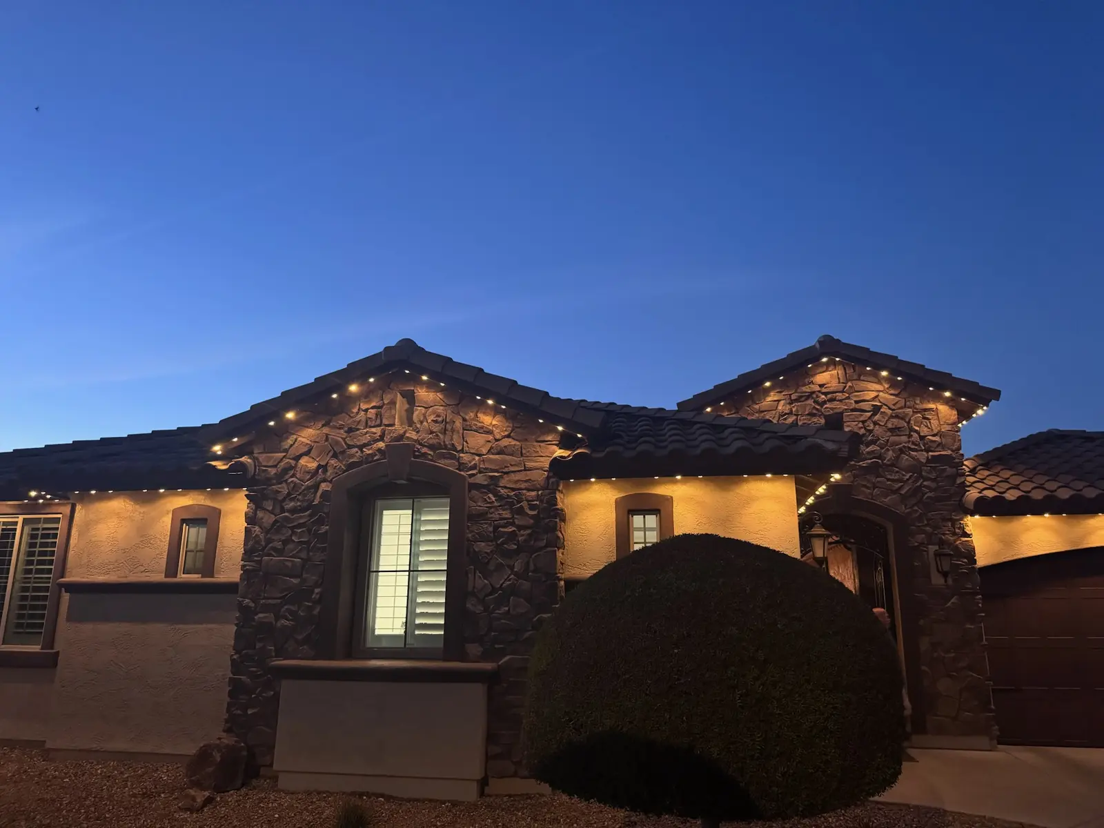 Stone front home lit in warm white permanent LED lighting along the roofline and entry arch