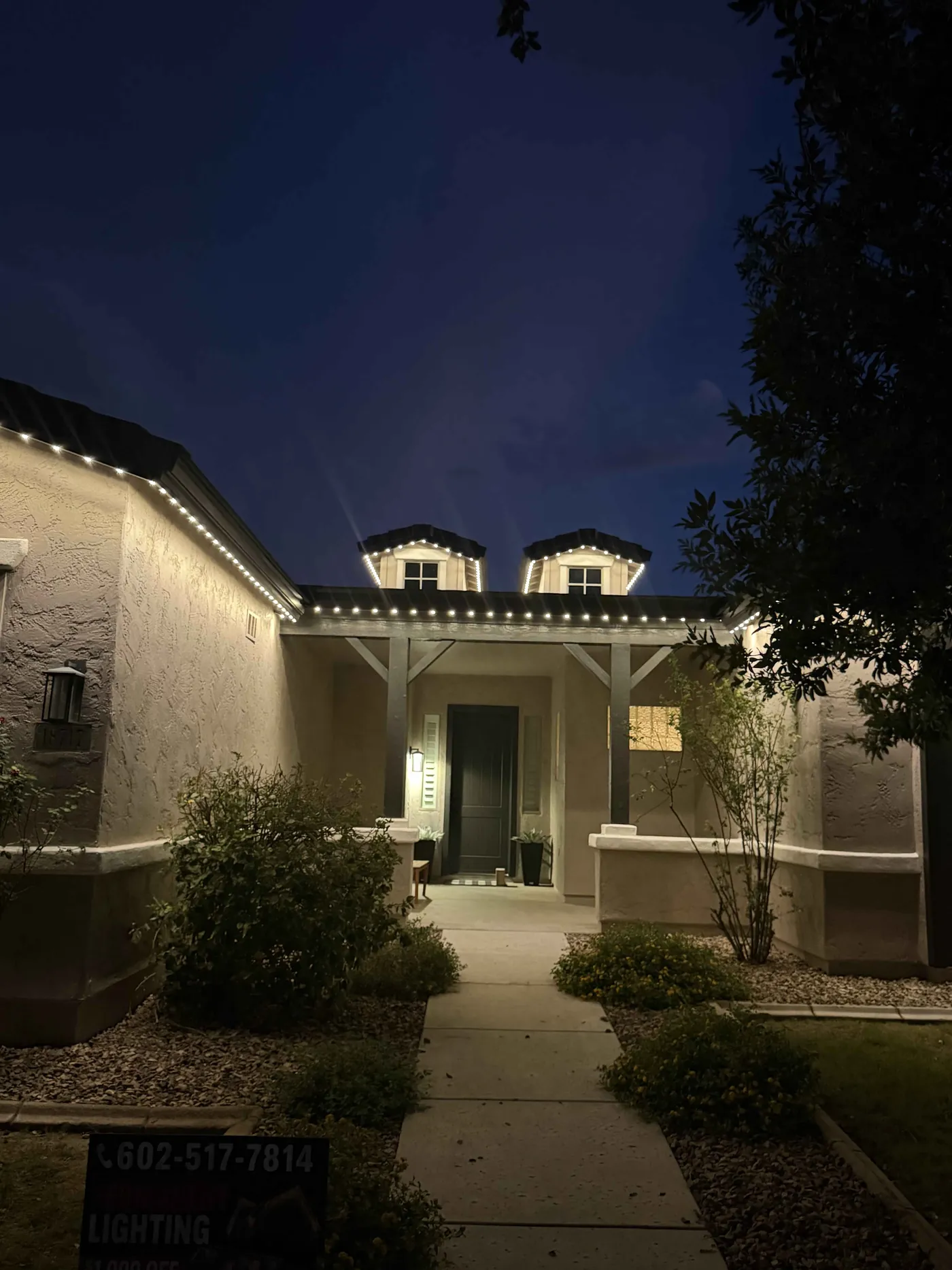 Single story home with warm white permanent lighting on the roofline and dormers, a Red Rock Lighting yard sign out front